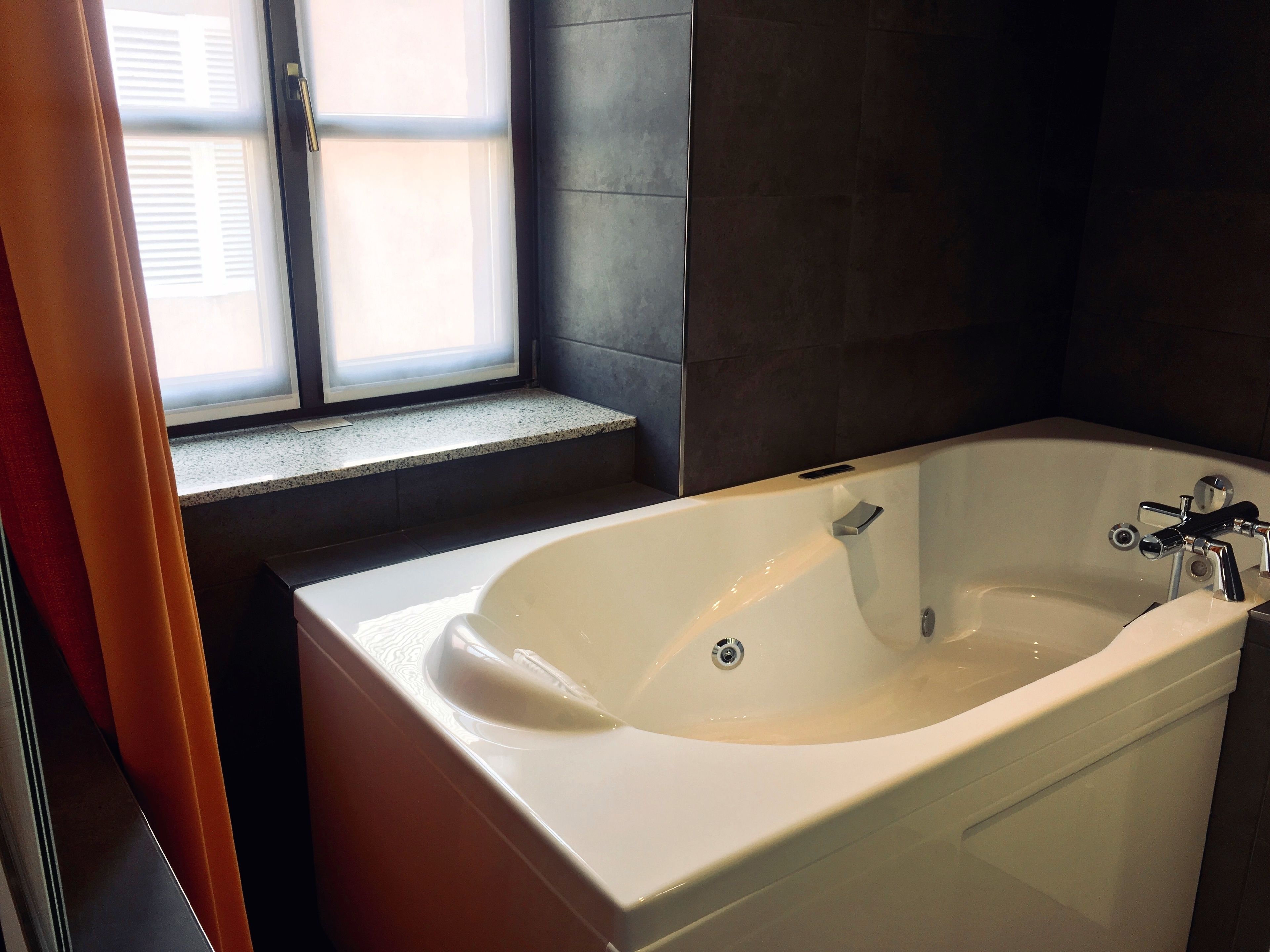 A modern bathroom with a freestanding bathtub, a window with blinds, and dark tiled walls.