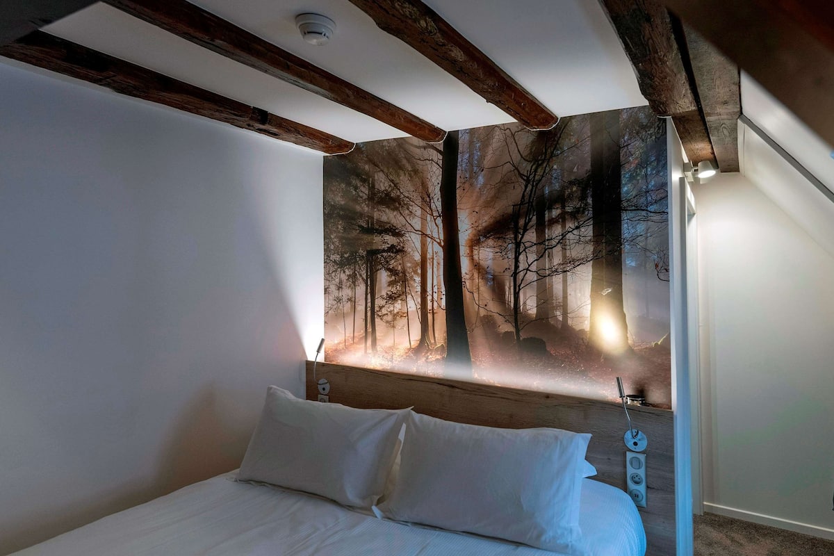 Un dormitorio con cama, almohadas blancas, cabecera con un mural de bosque y techo con vigas de madera a la vista.
