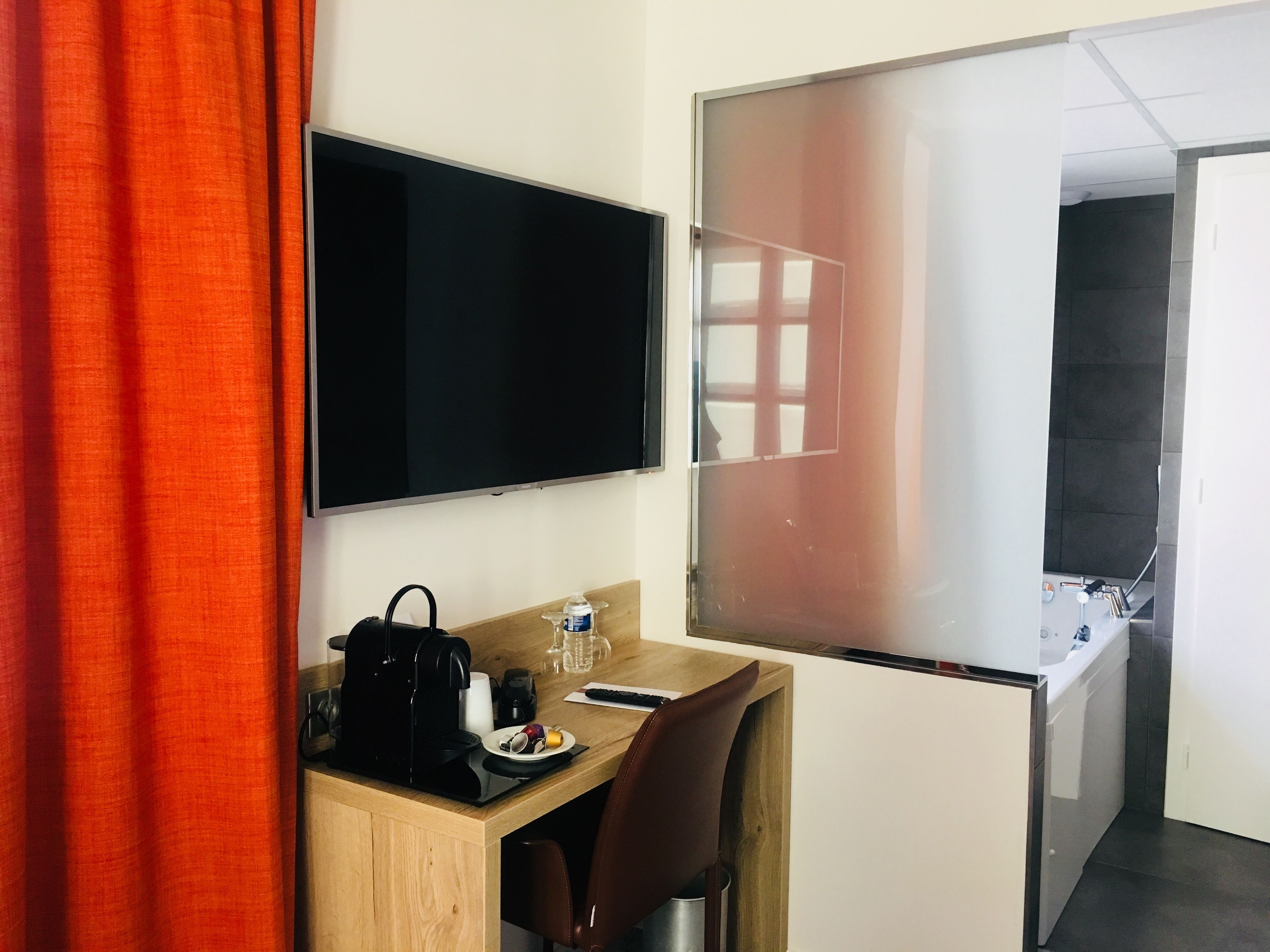 Une chambre d’hôtel avec un bureau en bois, une machine à café noire, une assiette avec des fruits, une bouteille d’eau, une chaise marron, une télévision et une porte en verre dépoli donnant sur une salle de bain.