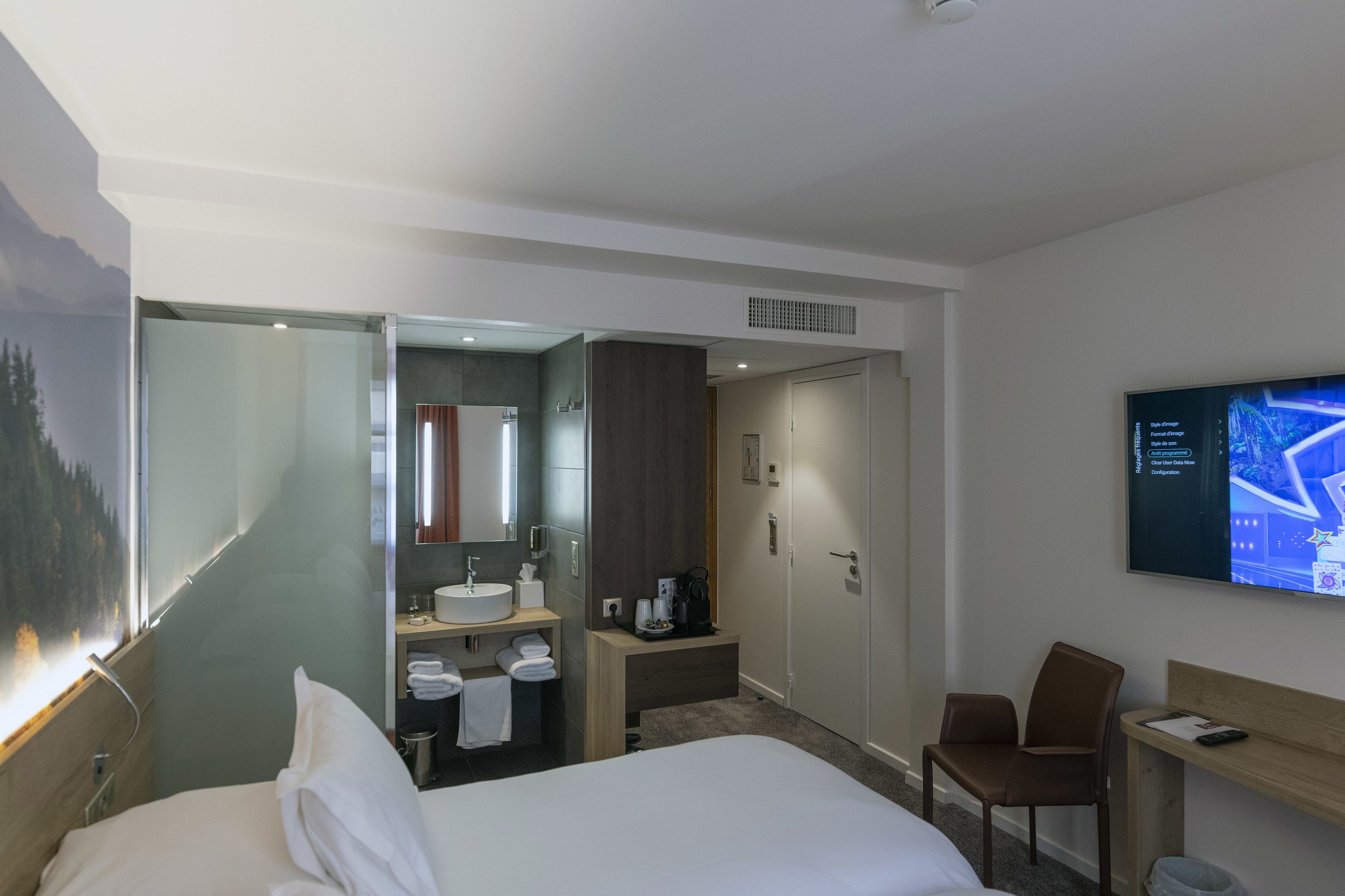 Hotelkamer met een bed, een bureau, een stoel, een televisie, een badkamer met een wastafel en spiegel, en een tv aan de muur.