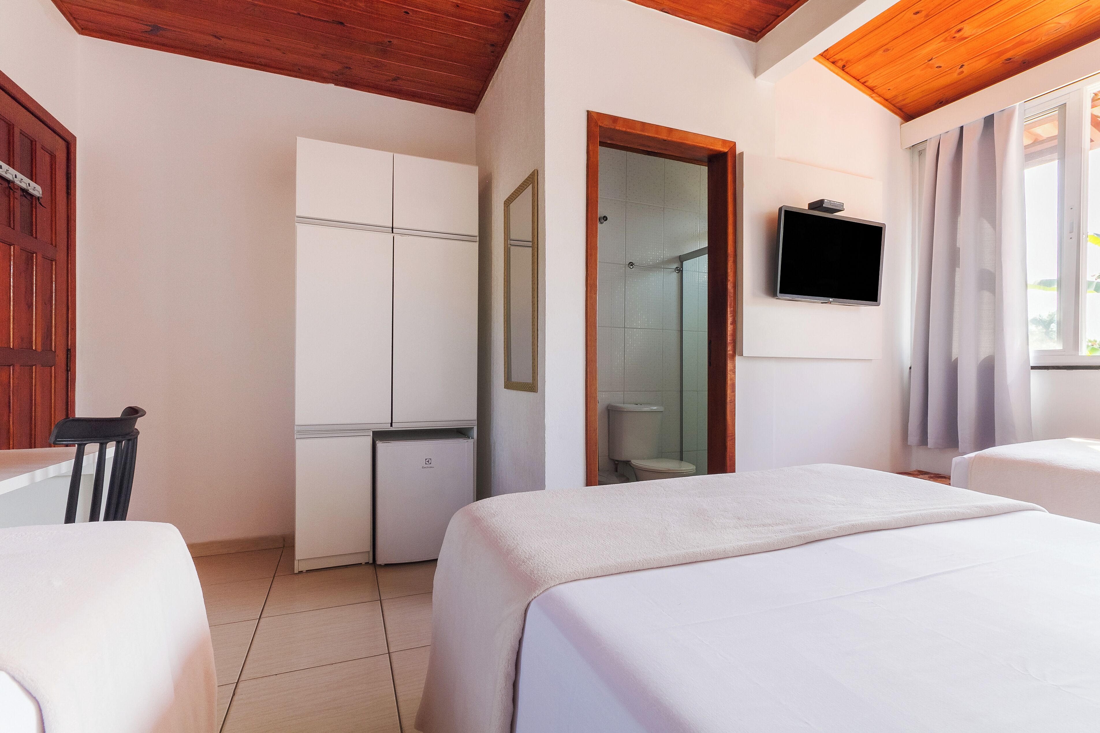 A hotel room with a bed, a desk, a small refrigerator, a mirror, a television, and a shower area.