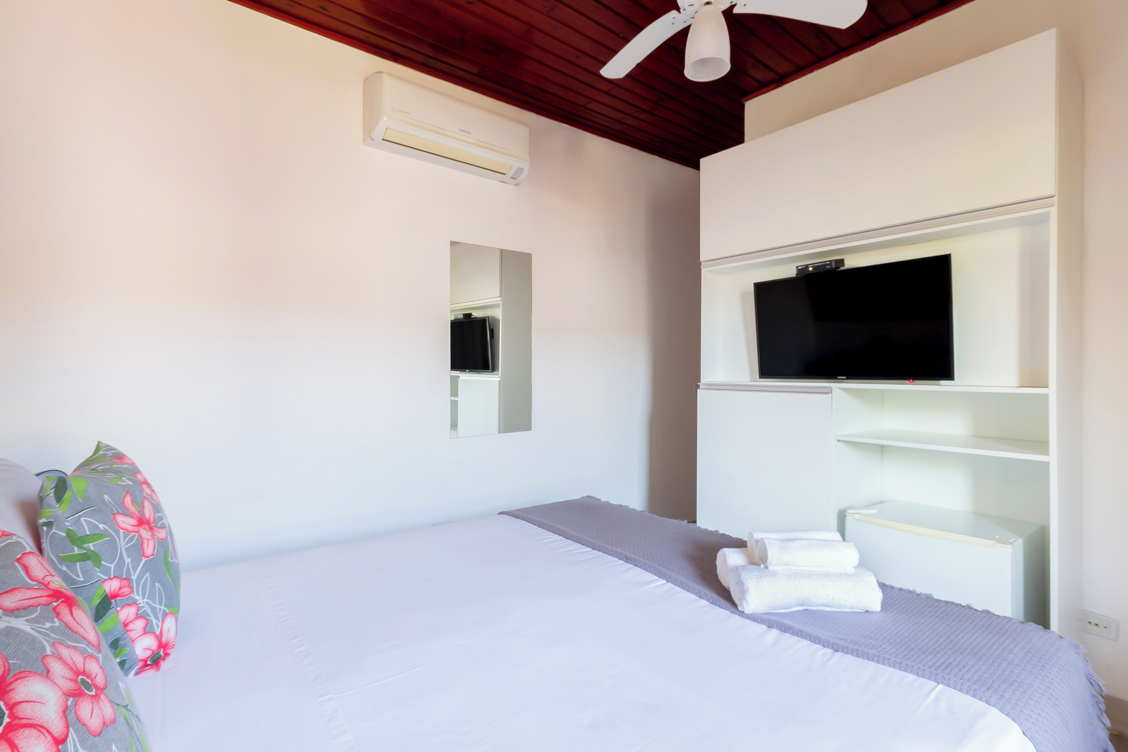A bedroom with a bed, a TV mounted on a white cabinet, a ceiling fan, and an air conditioning unit.