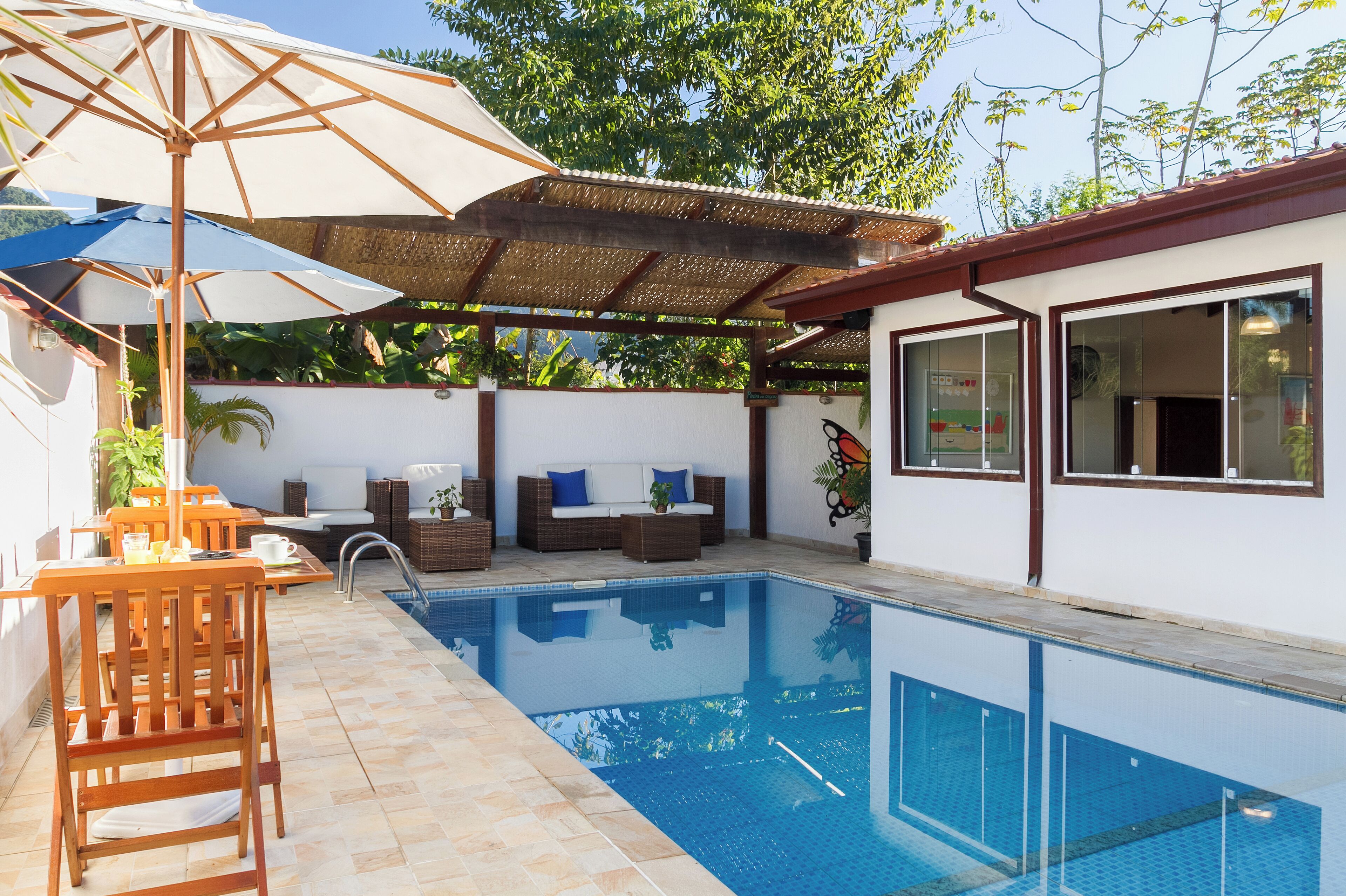 Una piscina con una sombrilla, sillas de madera y un sofá bajo una pérgola.