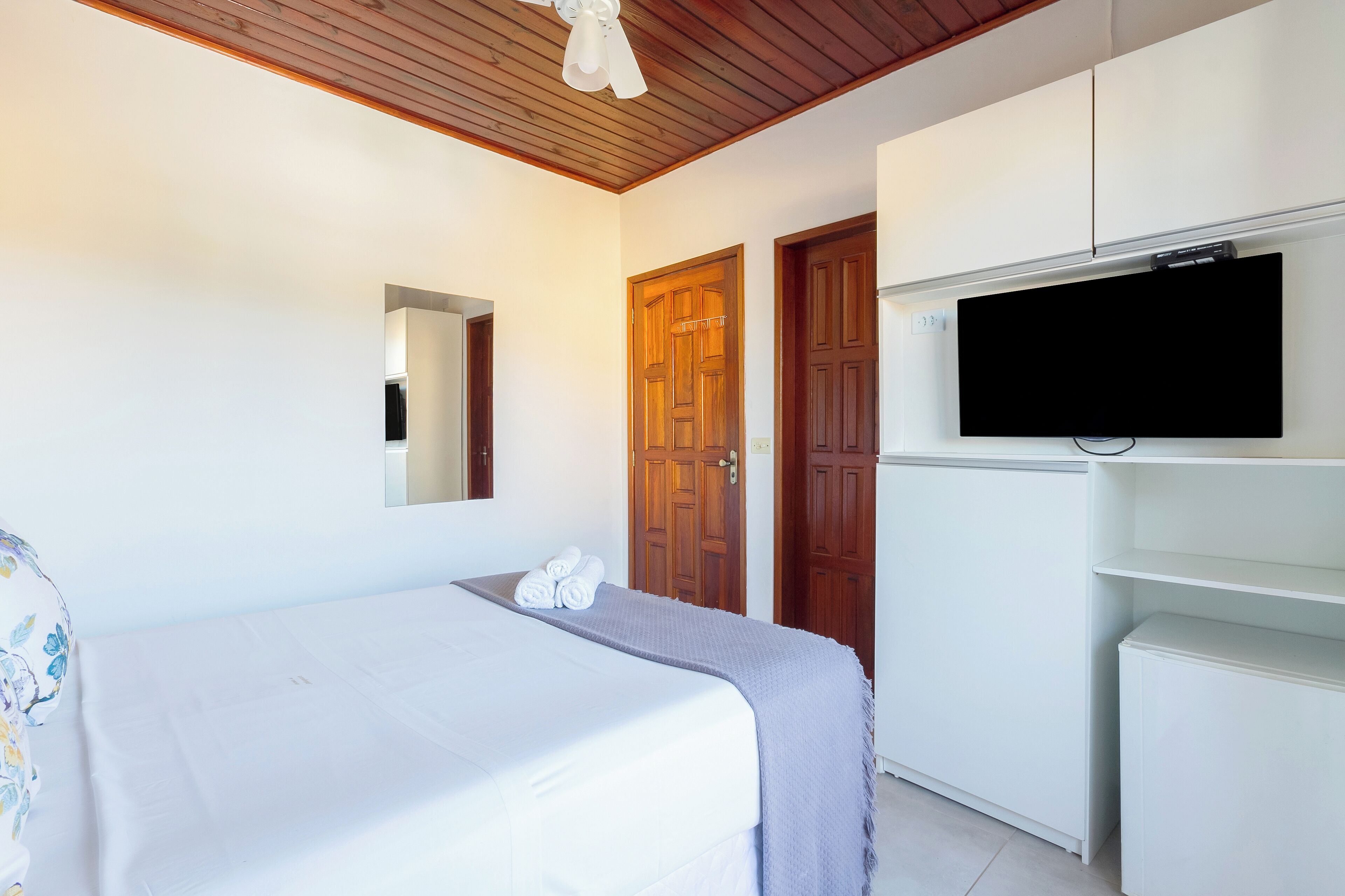 Quarto com cama, televisão na parede, portas de madeira e ventilador de teto.