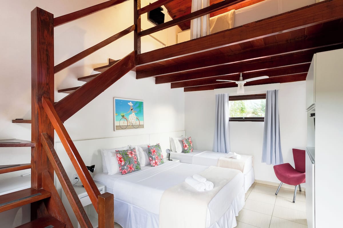 A hotel room with a wooden staircase, two beds, a red chair, and a window with curtains.