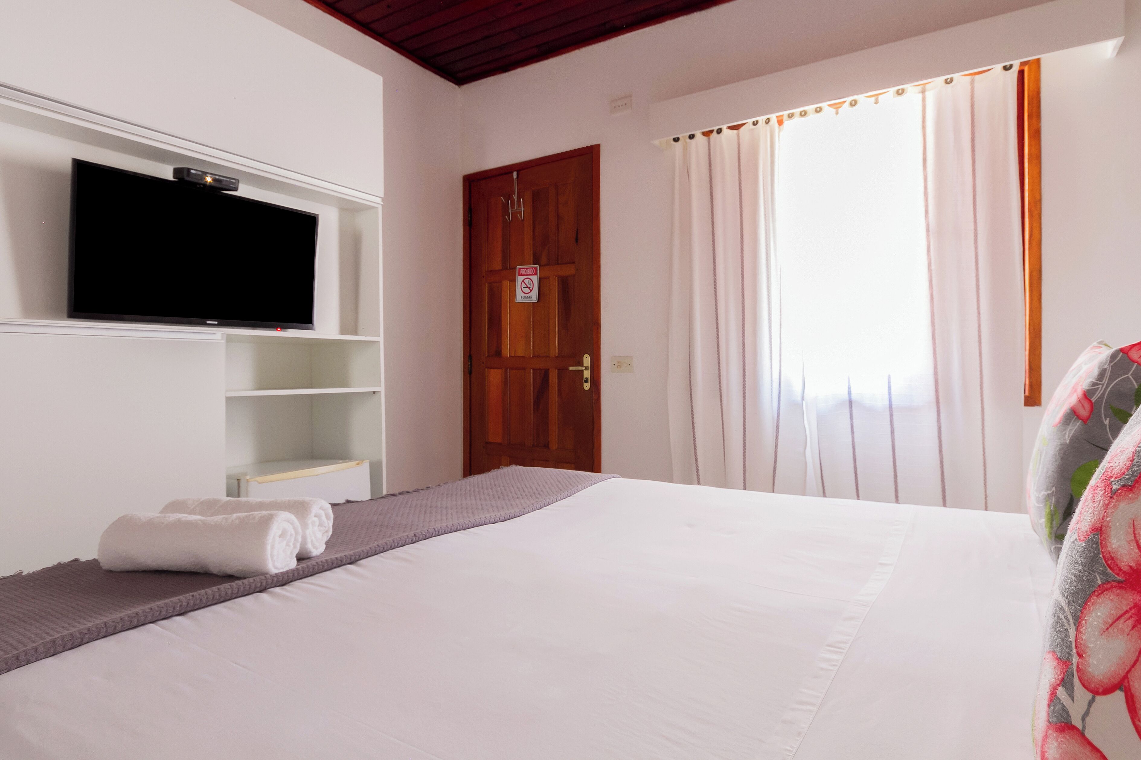 A hotel room with a bed, a television mounted on the wall, a wooden door, and a window with curtains.
