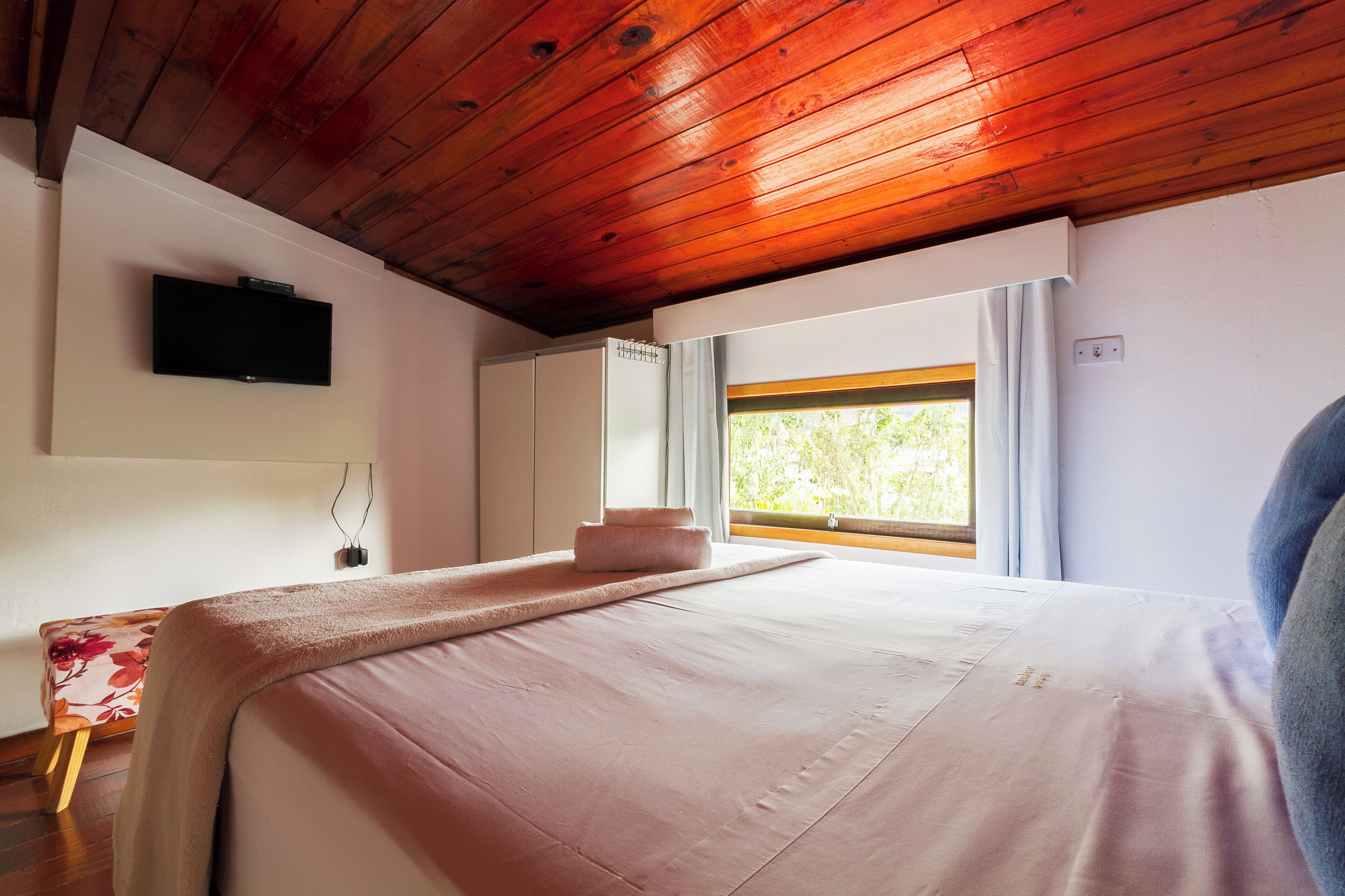 Un dormitorio con techo de madera, una cama con colcha rosa, un televisor de pantalla plana en la pared y una ventana con cortinas.