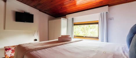 Un dormitorio con techo de madera, una cama con colcha rosa, un televisor de pantalla plana en la pared y una ventana con cortinas.