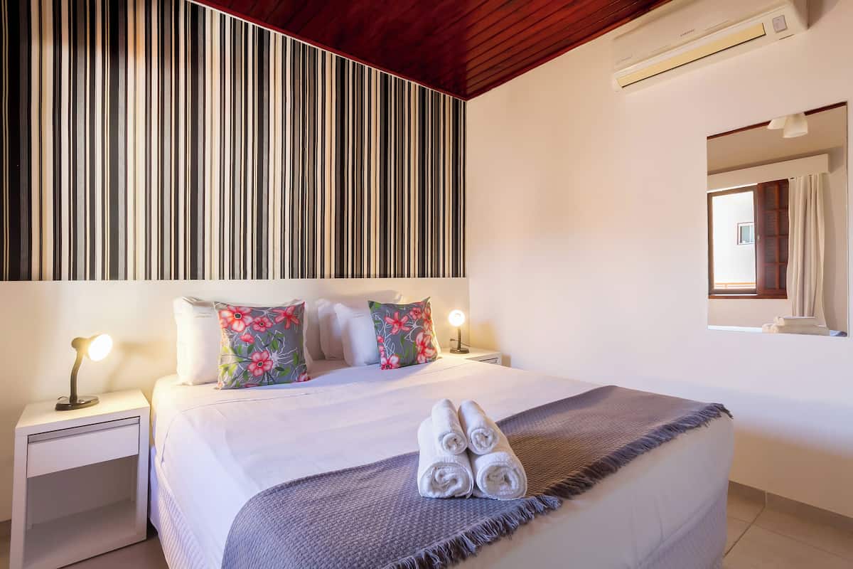 A hotel room with a large bed, two bedside tables, a wall-mounted air conditioner, and a window with curtains.