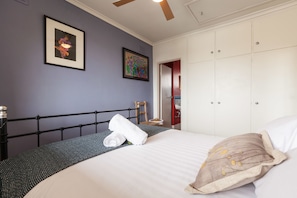 Een netjes opgemaakt bed met een kussen en een opgerolde handdoek, een lijst met kunstwerk aan de muur, en een garderobe met spiegel.
