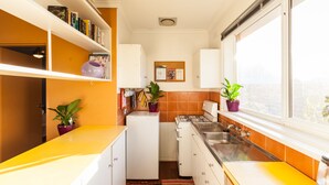 Une cuisine de taille réduite avec des meubles blancs, une cuisinière et un évier. On y trouve deux plantes en pot sur le comptoir et une fenêtre donnant sur l’extérieur.