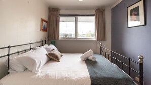 Un lit bien fait, avec des draps blancs, un plaid vert foncé et une serviette pliée. Il y a une fenêtre avec des rideaux, un ventilateur de plafond et un tableau encadré au mur.