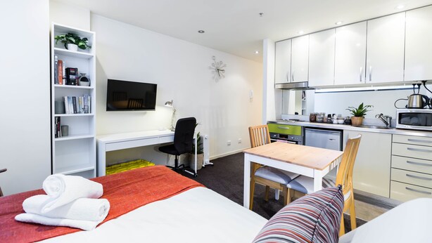 Una habitación de hotel moderna con cama, escritorio, silla, televisor, estantería y kitchenette.