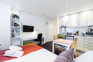 A modern hotel room with a bed, a desk, a chair, a TV, a bookshelf, and a kitchenette. - EMILY, Carlton Studio Apartment (Carlton)