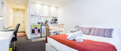A modern hotel room with a bed, a desk, a chair, and a kitchenette.