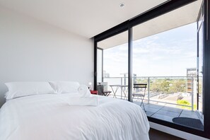 A modern bedroom with a large bed, a small table, and a balcony with a view of the city. - ERICA, 2BDR South Yarra Apartment (South Yarra)