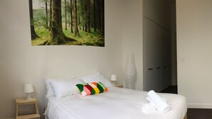 Ein Hotelzimmer mit Bett, weißer Bettwäsche, einem farbenfrohen Kissen, einem Nachttisch mit Lampe, einem großen Wandposter mit Waldmotiv und einem Kleiderschrank.