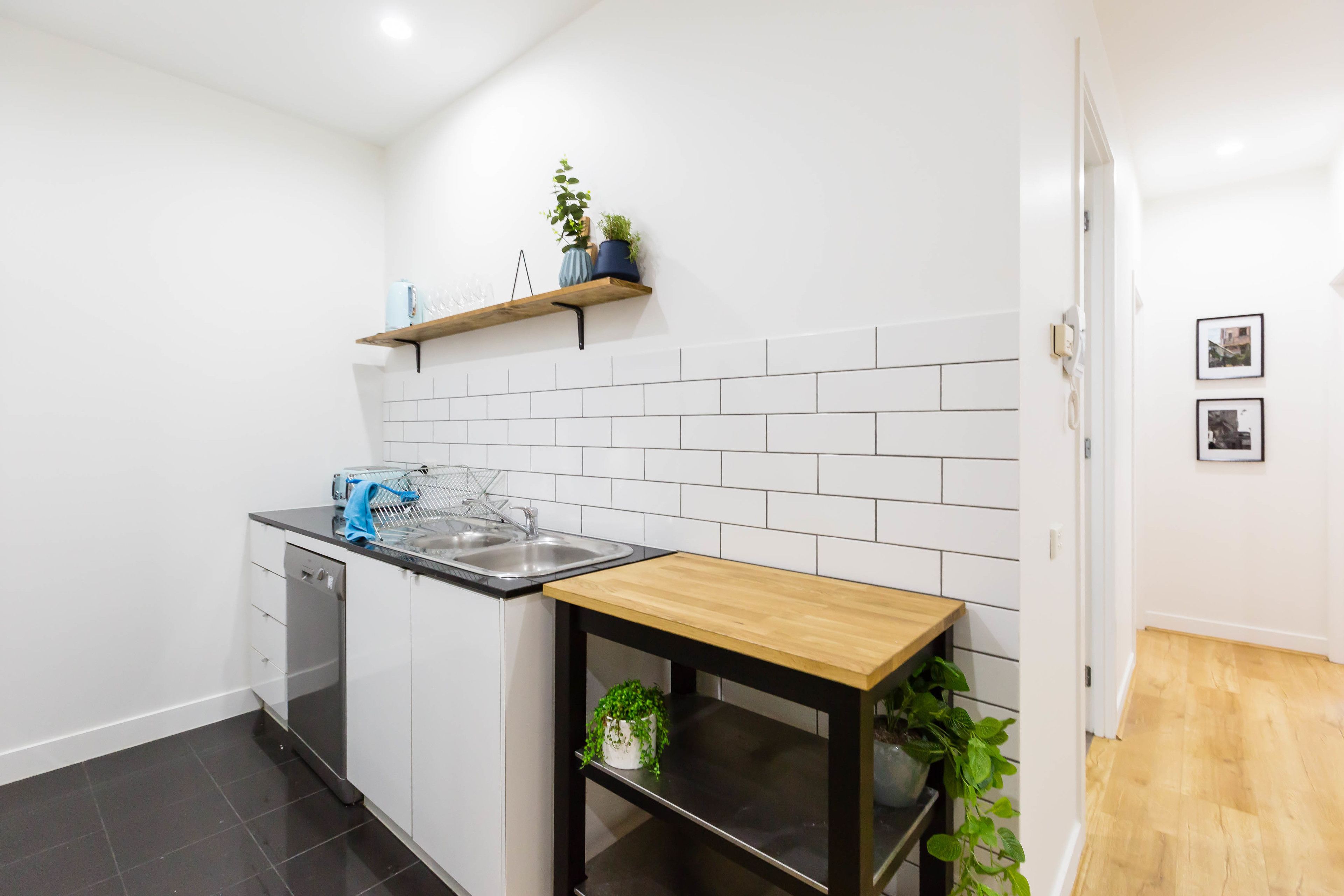 Uma cozinha moderna com armários brancos, bancada preta, mesa de madeira e prateleira suspensa com plantas.