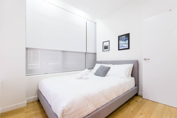 A modern bedroom with a large window, a bed with white bedding, a grey headboard, and a framed picture on the wall.