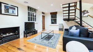 A modern living room with a fireplace, a black sofa, a TV, and a staircase. - HELENA, 2BDR Fitzroy House (Fitzroy)
