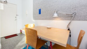 Una habitación con una mesa de madera, dos sillas, una lámpara de escritorio, un tablón de anuncios y una puerta con extintor.