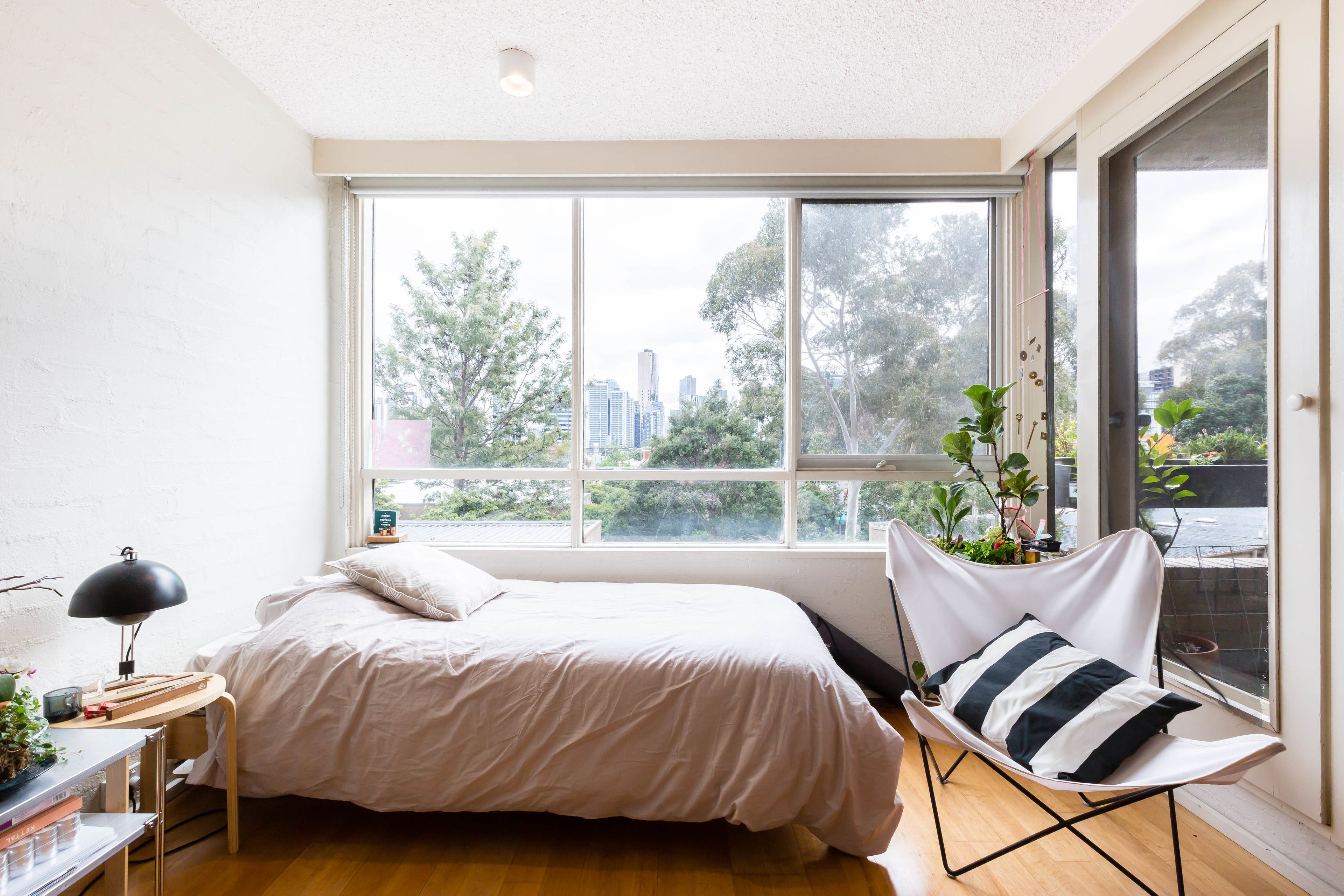 A bedroom with a large window, a bed, a desk with a lamp, and a chair with a plant.