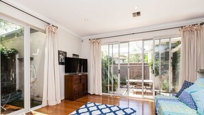 A spacious room with a large sliding glass door leading to a patio, a wooden dresser, a TV, and a patterned rug. - JASMINE, 2BDR Port Melbourne House (Port Melbourne)