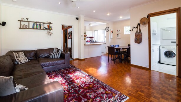 A spacious living area with a brown leather sofa, a red patterned rug, and a ceiling fan. - KELLY, 3BDR Northcote House (Northcote)