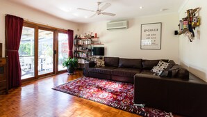 A living room with a brown sofa, a red patterned rug, a ceiling fan, and a bookshelf. - KELLY, 3BDR Northcote House (Northcote)