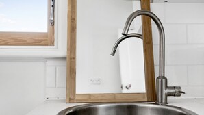 Un lavamanos de acero inoxidable con un grifo moderno, una ventana con marco de madera y paredes con azulejos blancos.