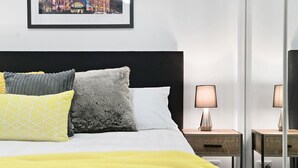 Una cama cuidadosamente tendida con una manta blanca y amarilla, un cabecero negro y cojines decorativos. Hay dos mesitas de noche con lámparas y una fotografía enmarcada en la pared sobre la cama.