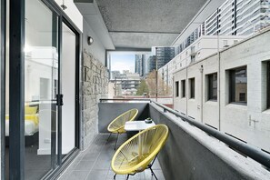 A balcony with two yellow chairs and a small table, overlooking a cityscape with modern buildings. - LIZZI, Melbourne Studio Apartment (Melbourne)