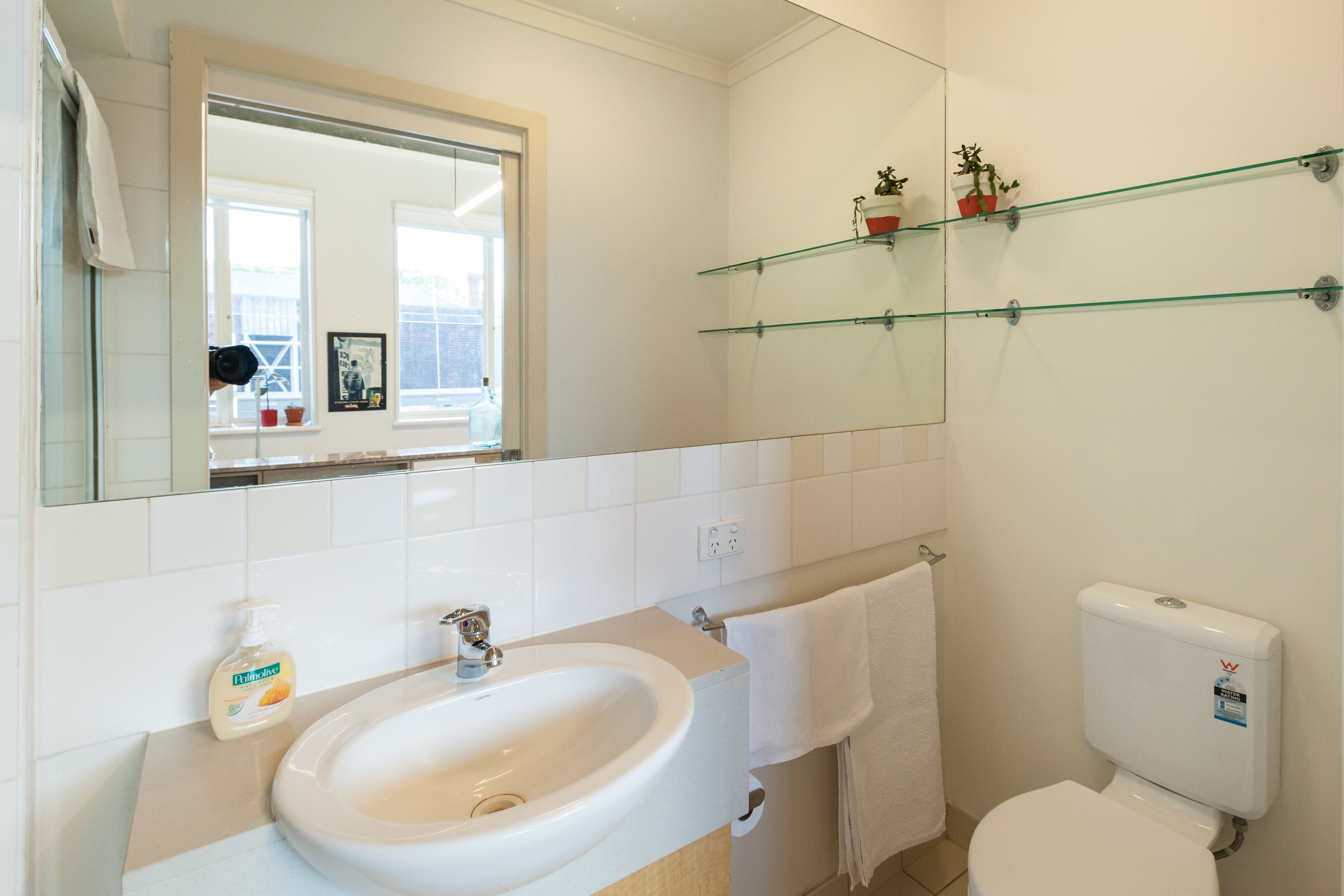 A bathroom with a white sink, mirror, and toilet. There are glass shelves above the toilet and a window with a view.