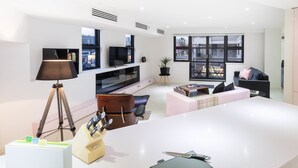 A modern living room with a large flat-screen TV, a tripod lamp, a dining area with a white table, and a sofa with a pink cushion. - MADDISON, 3BDR Port Melbourne Apartment (Port Melbourne)