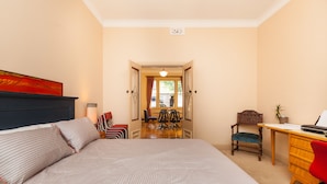 A bedroom with a bed, a desk, a chair, and a view of the dining area through an open door.