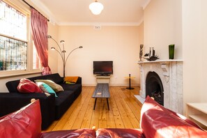 A living room with a black sofa, a wooden coffee table, a flat-screen TV, a fireplace, and a large window with curtains. - MADELINE, 1BDR Fitzroy Apartment (Fitzroy)