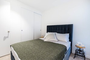 A neatly made bed with a dark headboard, a green blanket, and white pillows. There is a small wooden side table with a tissue box beside the bed. - MAXINE, 1BDR Collingwood Apartment (Collingwood)
