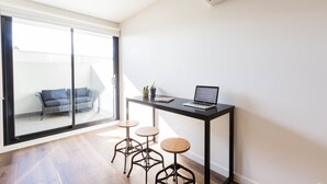 Una habitación moderna con una puerta de cristal corredera que da a un balcón, un escritorio pequeño negro con una computadora portátil y dos taburetes.