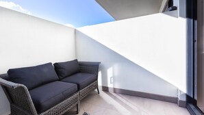 Un balcón con un sofá de mimbre y una mesita pequeña, una pared blanca y un cielo despejado.