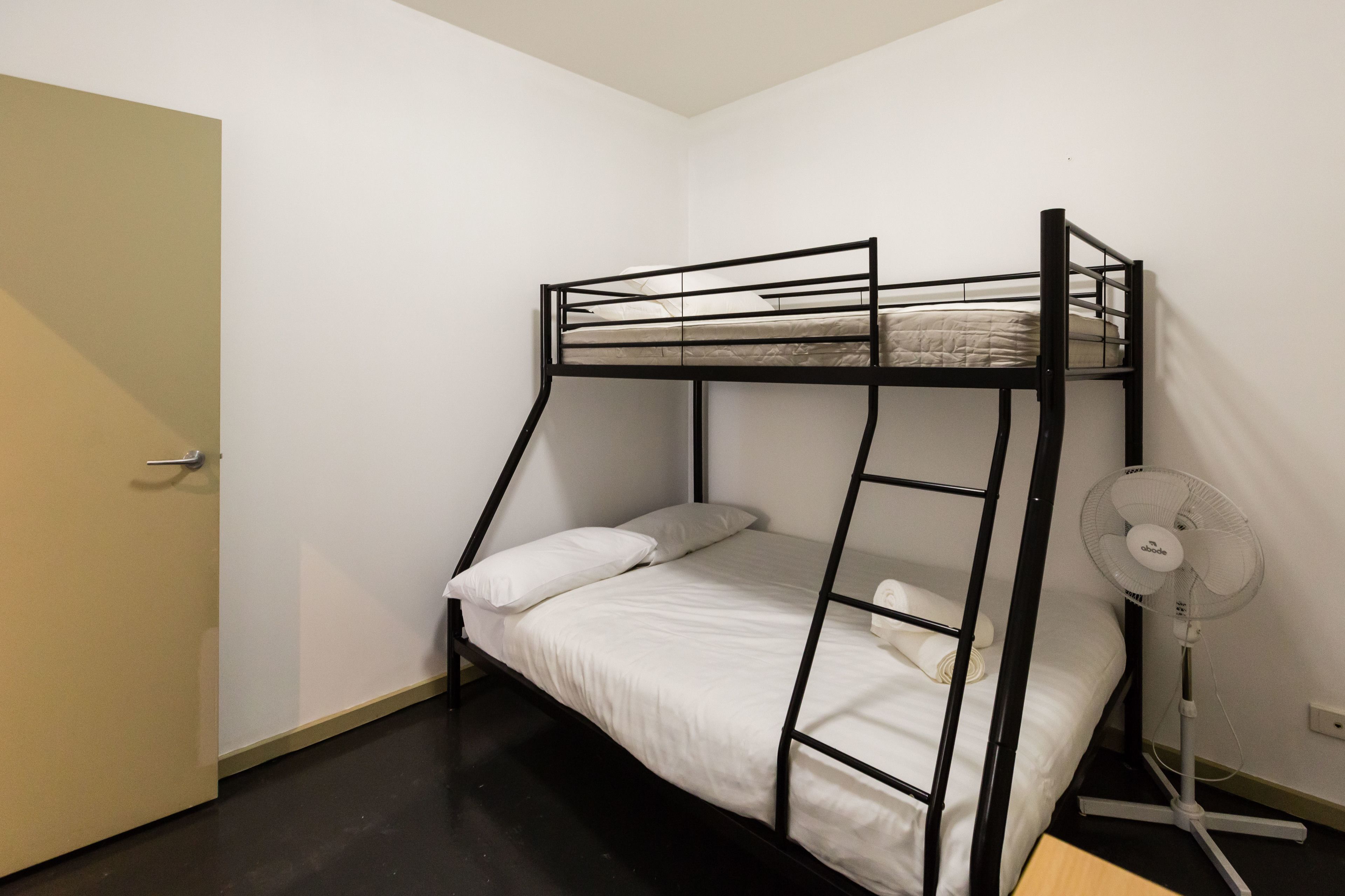 A bunk bed with white bedding, a standing fan, and a door to the left.