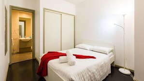 Habitación de hotel con cama, mesita de noche, manta roja, toalla blanca, lámpara de pie y vista al baño.