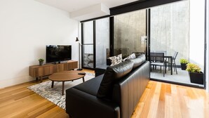 A modern living room with a black leather sofa, a wooden coffee table, and a flat-screen TV on a wooden cabinet. Large glass doors lead to an outdoor patio with a dining table and chairs. - RENEE, 4BDR North Melbourne House (North Melbourne)