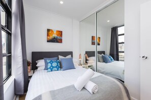 Habitación de hotel moderna con una cama grande, vistas a la ciudad, un cuadro en la pared y un espejo que refleja la habitación.