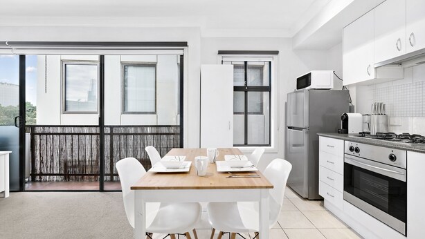 A modern kitchen with white cabinets, a stainless steel refrigerator, and a wooden dining table with white chairs. - SIENNA, 1BDR South Melbourne Apartment (South Melbourne)