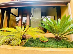 Un edificio con vestíbulo cubierto, plantas de palma y un pequeño elemento acuático.