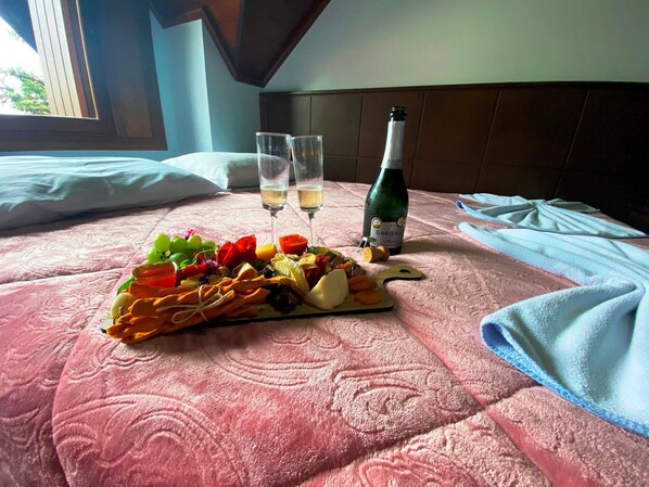 Una cama con un edredón rosa, una botella de vino, dos copas y una tabla de fiambres sobre la cama.