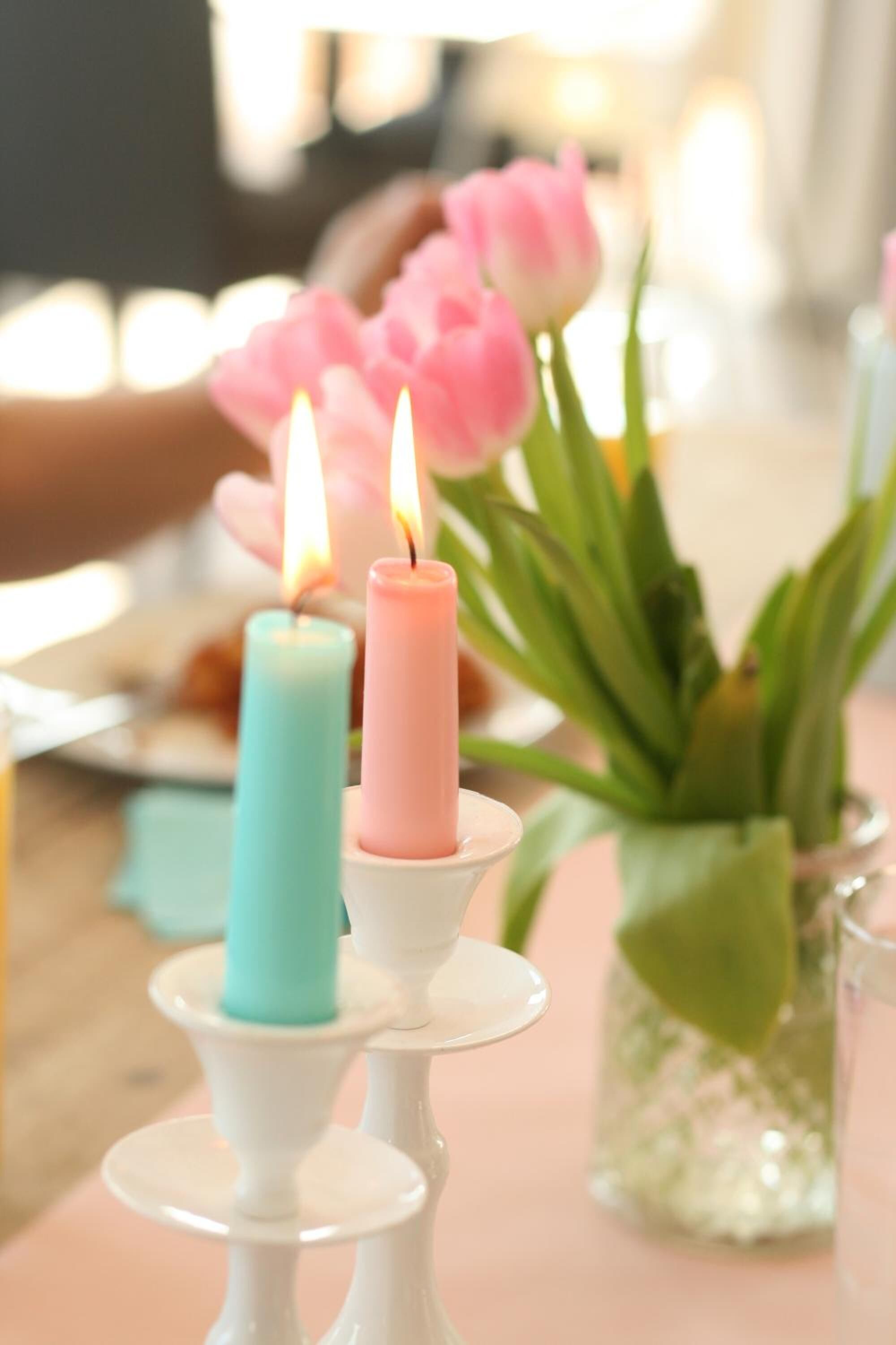 A table with two lit candles on white stands, a vase with green leaves, and pink flowers in the background.