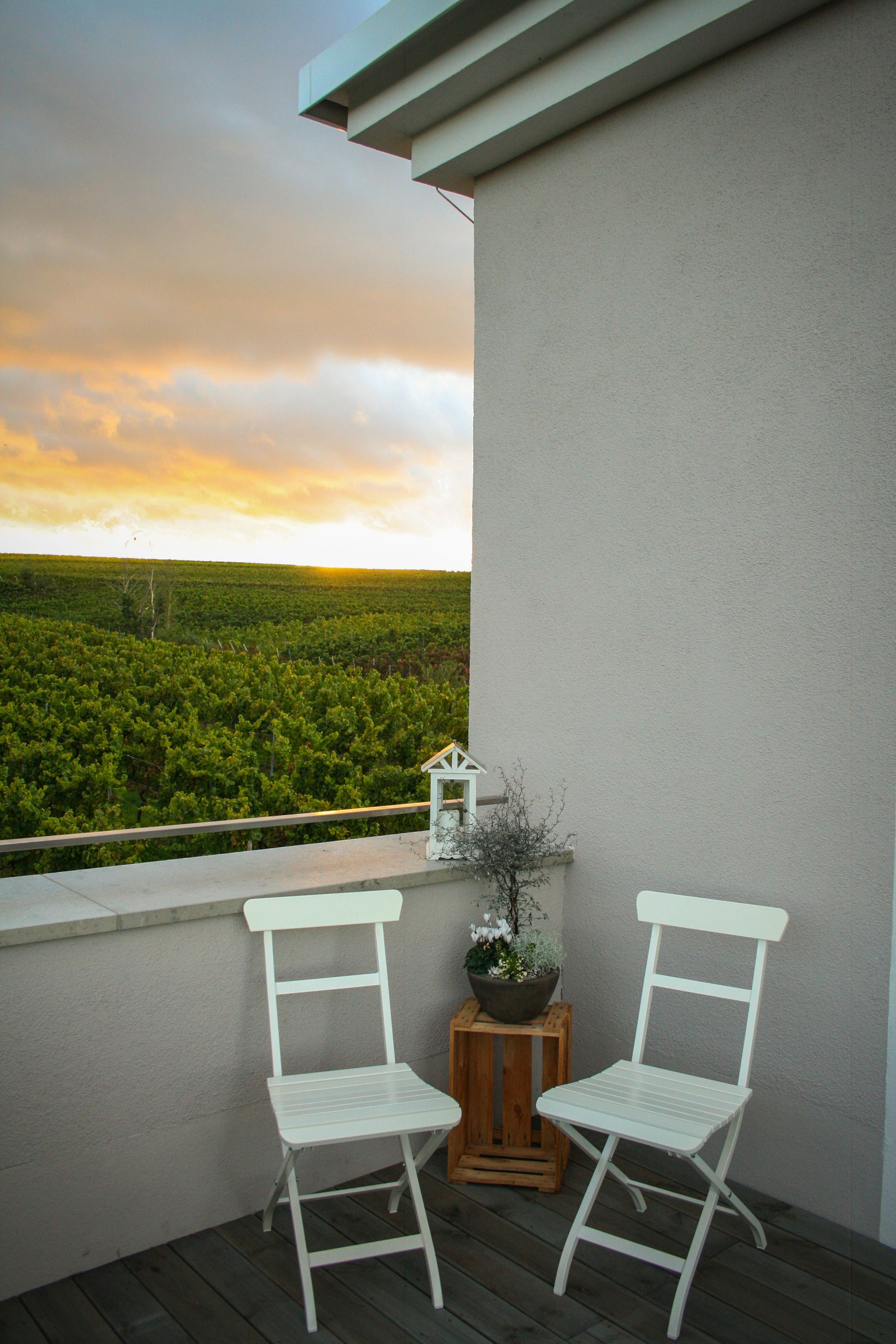 A balcony with two white chairs, a small wooden table with a potted plant, and a view of a green landscape under a cloudy sky.