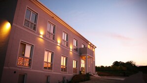 A two-story building with multiple windows and balconies, featuring a well-lit entrance and a driveway leading to the front door. - Trautwein - Das Winzerhotel am La Roche (Flonheim)