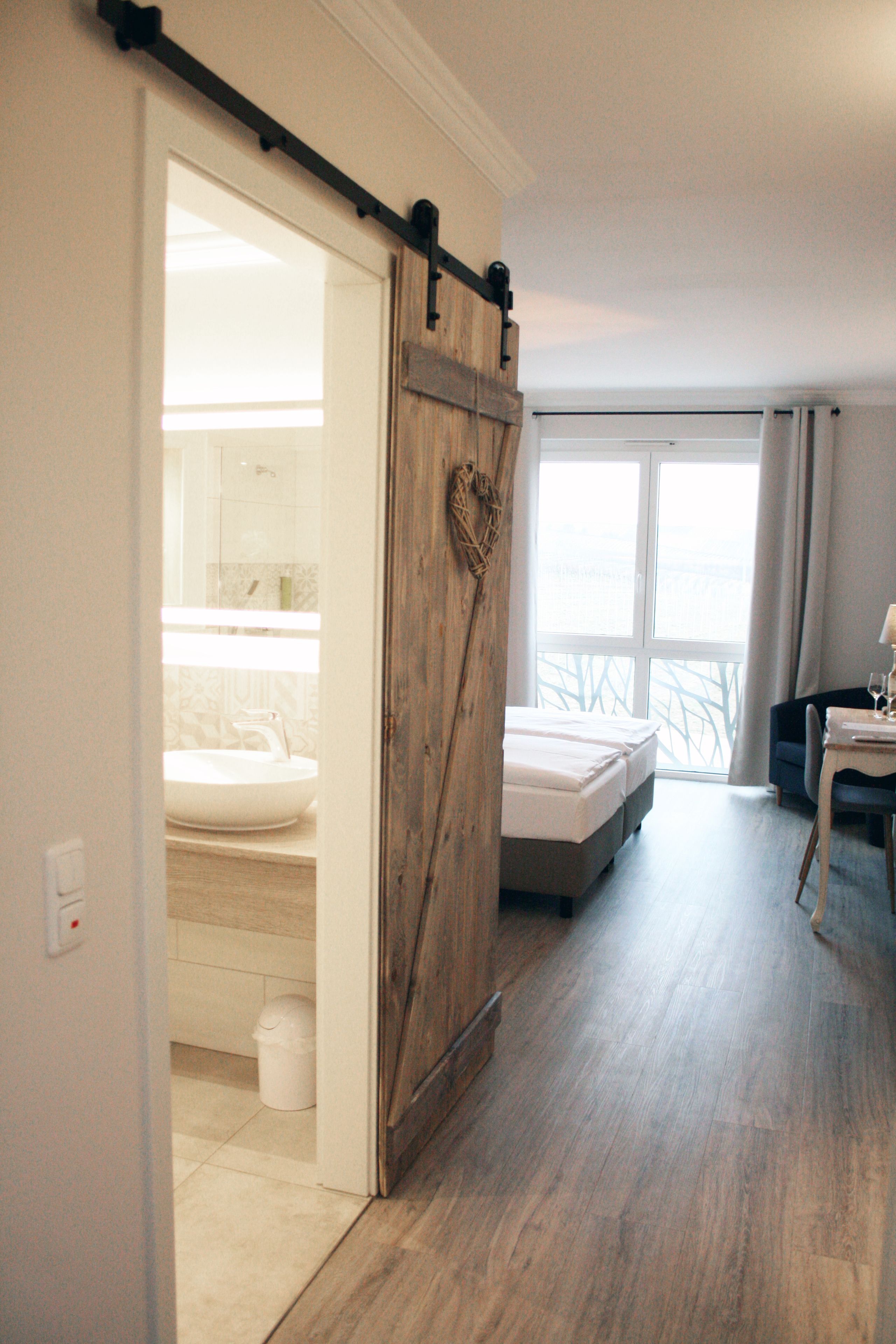 Quarto de hotel com porta de madeira deslizante, cama, escrivaninha com cadeira e vista para o exterior.